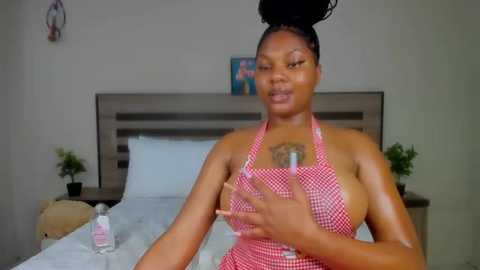 Media: Video of a dark-skinned Black woman with braided hair, wearing a red-and-white checkered apron, sitting on a bed in a minimalistic bedroom.