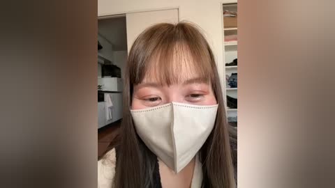 Media: Video of an Asian woman with long brown hair and bangs, wearing a beige face mask, standing in a modern kitchen with white cabinets and wooden floors.