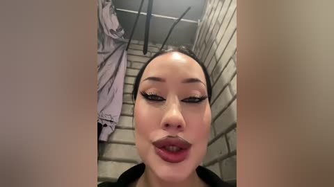 Media: Video of an Asian woman with light skin, high cheekbones, and full lips, wearing heavy black eyeliner, captured in a dimly-lit brick stall with a white jacket hanging on a hook.