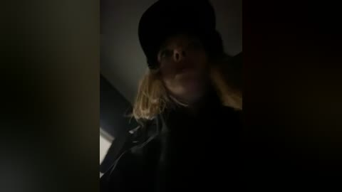 Media: A dimly lit video of a woman in a black hat and coat, with a hint of blonde hair visible, looking up with a serious expression.