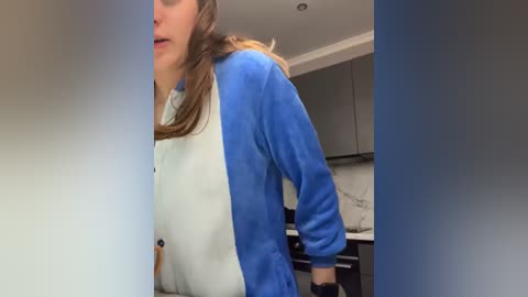 Media: A video shows a woman in a blue and white fuzzy jacket with a hood, standing in a modern kitchen with white cabinets and a marble backsplash.