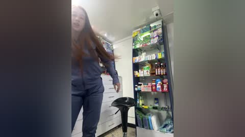 Media: Video of a tall, slender woman with long, straight brown hair, wearing a dark blue jumpsuit, entering a modern kitchen with white cabinets and a tall, brightly lit pantry filled with colorful groceries.