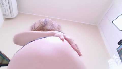 Media: A low-angle video of a naked, light-skinned woman with long blonde hair, bending over, her hand on her buttocks, in a minimalist room with a window and air conditioner.