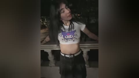 Media: A video of a young woman with long black hair, wearing a cropped white T-shirt with a purple cartoon character and black pants, standing indoors.