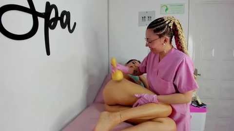 Media: Video of a woman with braided hair in pink scrubs holding a yellow vibrator over a woman's genitals in a white room with an \"Open\" sign.