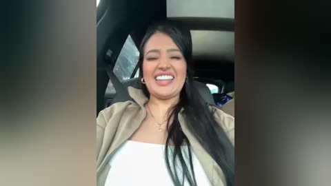 Media: Video of a young woman with long black hair, wearing a beige jacket over a white top, smiling widely in a car.