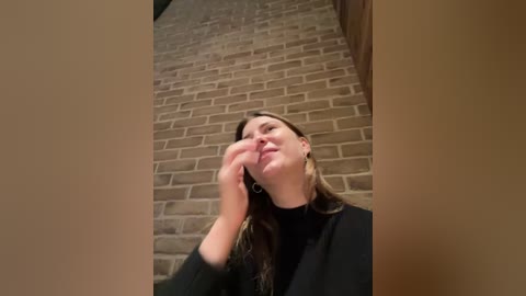 Media: Video of a young woman with long brown hair, wearing a black top, standing against a brick wall. She is touching her face, smiling, and appears to be in a playful or contemplative mood.