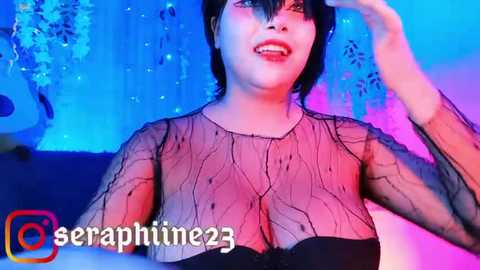 Media: Video of a smiling, fair-skinned woman with short black hair, wearing a sheer black top that reveals cleavage, in a colorful, sparkly room.