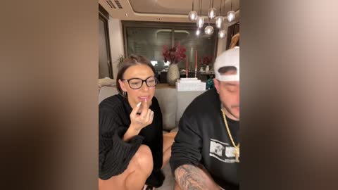 Media: A video of a young woman with glasses and short dark hair, wearing a black dress, eating a cookie, and a man in a black hoodie and white cap, sitting in a modern living room with beige furniture and a large window.