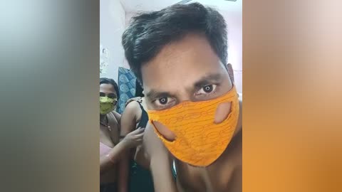 Media: Video of a young Indian man with short dark hair and light skin, wearing a bright orange mask, standing in a small, dimly lit room with a woman in the background, both in casual attire.