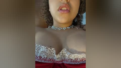 Media: Video of a woman with curly hair, wearing a lacy white and red strapless top, and a silver choker necklace. Her lips are slightly parted, showcasing her tongue.