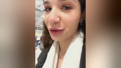 Media: A close-up video of a young woman with fair skin, brown hair, and light makeup, wearing a white and black jacket. She smiles slightly, with blurred store shelves in the background.