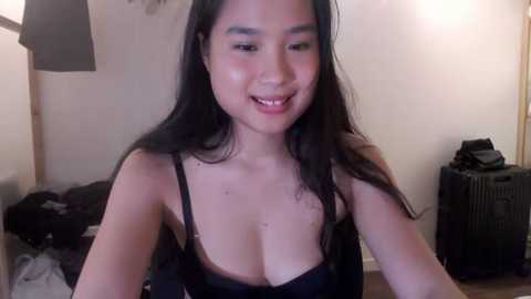 Media: Video of an Asian woman with long black hair, wearing a black bra, smiling. Background includes a bed, a lamp, and a black suitcase.