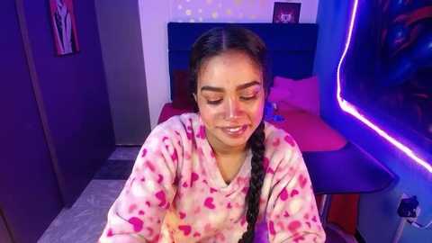 Media: Video of a young woman with medium skin tone, braided hair, and pink heart pajamas, smiling in a modern, dimly lit bedroom with a purple neon sign, red bed, and white wall with yellow polka dots.