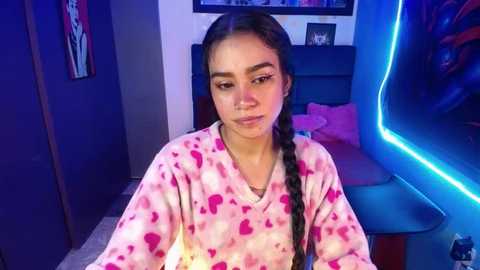 Media: Video of a young Latina woman with dark hair in a braid, wearing a pink, heart-patterned pajama set, sitting indoors in a dimly lit room with neon blue lighting.