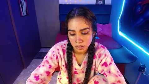 Media: Video of a young Latina woman with long black hair in twin braids, wearing a pink heart-patterned pajama set, sitting in a modern bedroom with blue neon lights.