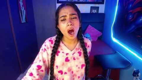 Media: Video of a young Latina woman with long braids, wearing pink pajamas with heart patterns, screaming in a gaming room with a neon blue light.