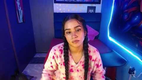 Media: Video of a young Latina girl with long braids in a pink tie-dye pajama set, sitting on a bed with a blue neon light strip.