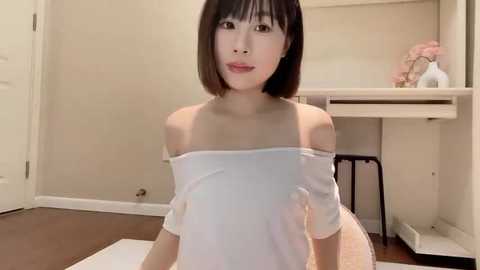 Media: Video of an East Asian woman with straight black hair and fair skin, wearing an off-shoulder white top, standing in a minimalist bedroom with beige walls, a vanity, and a pink flower vase.