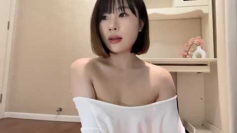 Media: Video of an Asian woman with short, straight black hair and fair skin, wearing an off-shoulder white top, sitting indoors with beige walls, a sink, and pink flowers in the background.