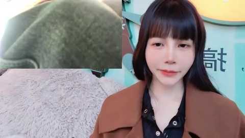 Media: Video of a young Asian woman with long black hair, wearing a brown coat over a black shirt, standing in a soft, light gray carpeted room with a blurred green fabric on the left.