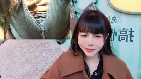 Media: Video of a fair-skinned East Asian woman with straight black hair, wearing a brown coat over a black shirt. Background shows partially visible denim-clad legs and a teal wall with Chinese characters.