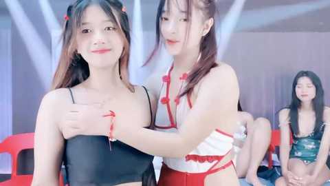 Media: Video of two young East Asian women in provocative, revealing outfits, one with a black crop top, the other in a white top with red trim, holding hands. Background shows a modern indoor setting with white and red decor.