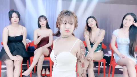 Media: Video of five Asian women in revealing outfits seated on red chairs under a bright stage light. The central woman has a short bob haircut and wears a white dress.