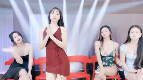 Media: Video of four Asian women, two in black dresses and two in red dresses, seated on bright orange plastic chairs, against a white backdrop, appearing poised and attentive.