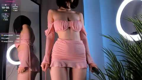 Media: A video of a slender, fair-skinned woman with short black hair, wearing a pink off-shoulder crop top and matching mini skirt, standing in front of a circular mirror with a glowing ring light.