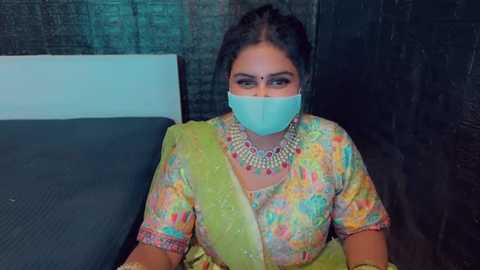 Media: Video of a South Asian woman in a colorful sari with intricate embroidery, wearing a blue surgical mask, seated on a dark couch in a dimly lit room with dark walls.