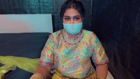 Media: Video of a woman in a colorful, patterned saree with a blue mask, seated on a bed, in a dimly lit room with dark, textured walls.