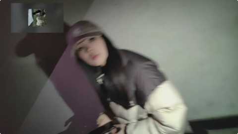 Media: A blurry video of a young Asian woman with long black hair and a gray hat, wearing a black jacket and beige pants, leaning against a wall with a shadowy figure in the background.