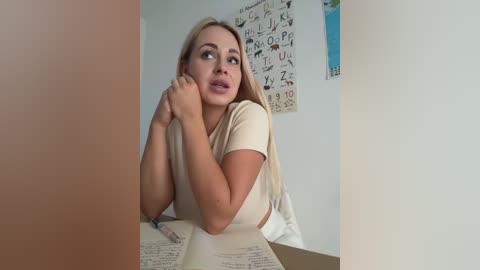 Media: A video of a blonde woman with a medium build and medium-sized breasts, wearing a beige t-shirt, pensively resting her chin on her hand while looking to the side. She sits at a cluttered desk with an open book and papers. The background features a white wall with educational posters.