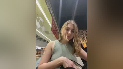 Media: Video of a young Caucasian woman with long blonde hair, wearing a sleeveless, light green top, standing in a dimly lit, modern subway station.