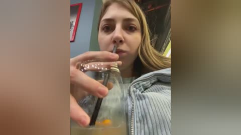 Media: A video shows a young woman with light skin and straight, shoulder-length brown hair, wearing a light gray hoodie. She is drinking from a glass bottle with a straw, set against a blurred background.