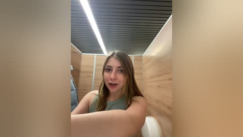 Media: Video of a young woman with straight brown hair, wearing a sleeveless green top, seated in a modern, wooden-paneled restroom. She looks directly at the camera with a neutral expression.