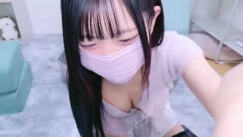 Media: Video of an Asian woman with long black hair, wearing a light pink mask and a sheer white top, kneeling on a grey floor in a modern, light-colored room.