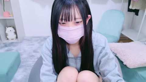 Media: Video of an Asian woman with long black hair, wearing a pink face mask, light blue hoodie, and sitting on a light blue chair in a softly lit, minimalist room.