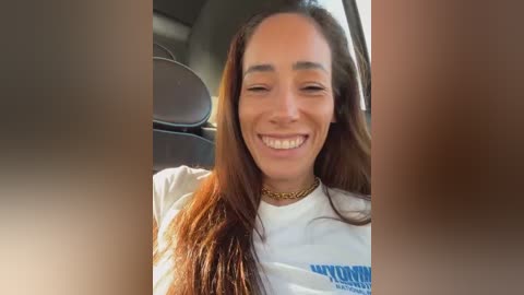 Media: Video of a joyful, light-skinned woman with long, straight brown hair, wearing a white T-shirt, smiling widely in a car.