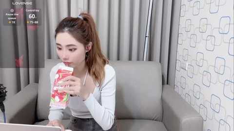 Media: Video of an Asian woman with a ponytail, wearing a white top and jeans, drinking from a red and white cup, seated on a gray sofa with a patterned wall behind her.