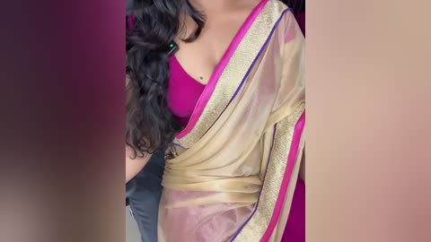 Media: Video of a woman's torso in a vibrant pink blouse, draped in a translucent beige sari with purple trim, against a blurred background. Her dark, curly hair cascades over her shoulder.