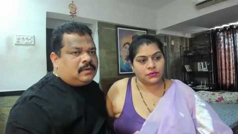 Media: Video of an Indian couple, man in black, woman in purple saree, in a modern living room with framed artwork and bookshelf.