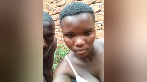 Media: Video of a young Black woman with short, curly hair and a medium complexion, wearing a white tank top. She gazes directly at the camera with a serious expression. The background features a brick wall and green foliage.