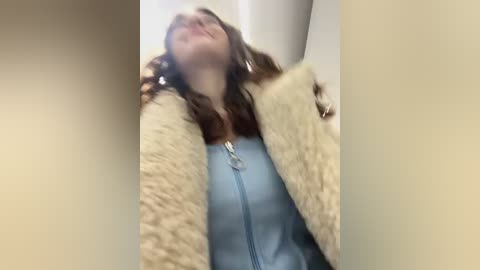 Video of a young woman with long brown hair, wearing a light blue shirt and a fluffy beige coat, standing indoors with a slightly blurred background.