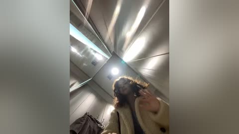 A video of a woman with shoulder-length, wavy hair, wearing a beige fur coat, walking down a metallic, sloped escalator. The background features light fixtures and blurred, reflective surfaces.