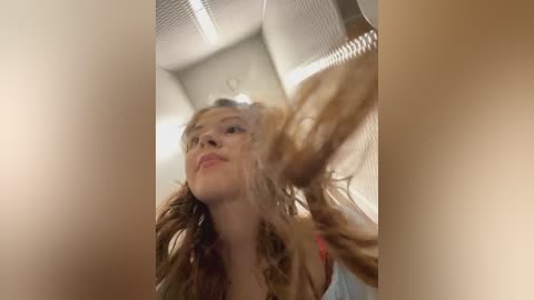 Video of a young woman with long, wavy blonde hair, captured in mid-action as she styles her hair. She has fair skin and wears a light blue top. The background features a modern, light-colored ceiling with recessed lighting.