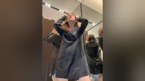 A video captures a woman in a black glittery dress with long sleeves, posing confidently in a mirrored room. She has shoulder-length blonde hair, and her arms are raised, hands behind her head.