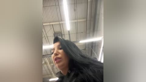 Media: Video of a woman with long black hair, wearing a black coat, captured from a low angle, in a dimly lit, industrial setting with a metal ceiling and fluorescent lights.