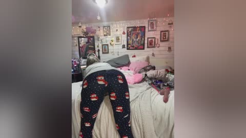 Media: Video of a blonde woman in pajamas, kneeling on a messy bed with scattered toys, in a cozy, cluttered bedroom with white brick walls adorned with framed art and fairy lights.